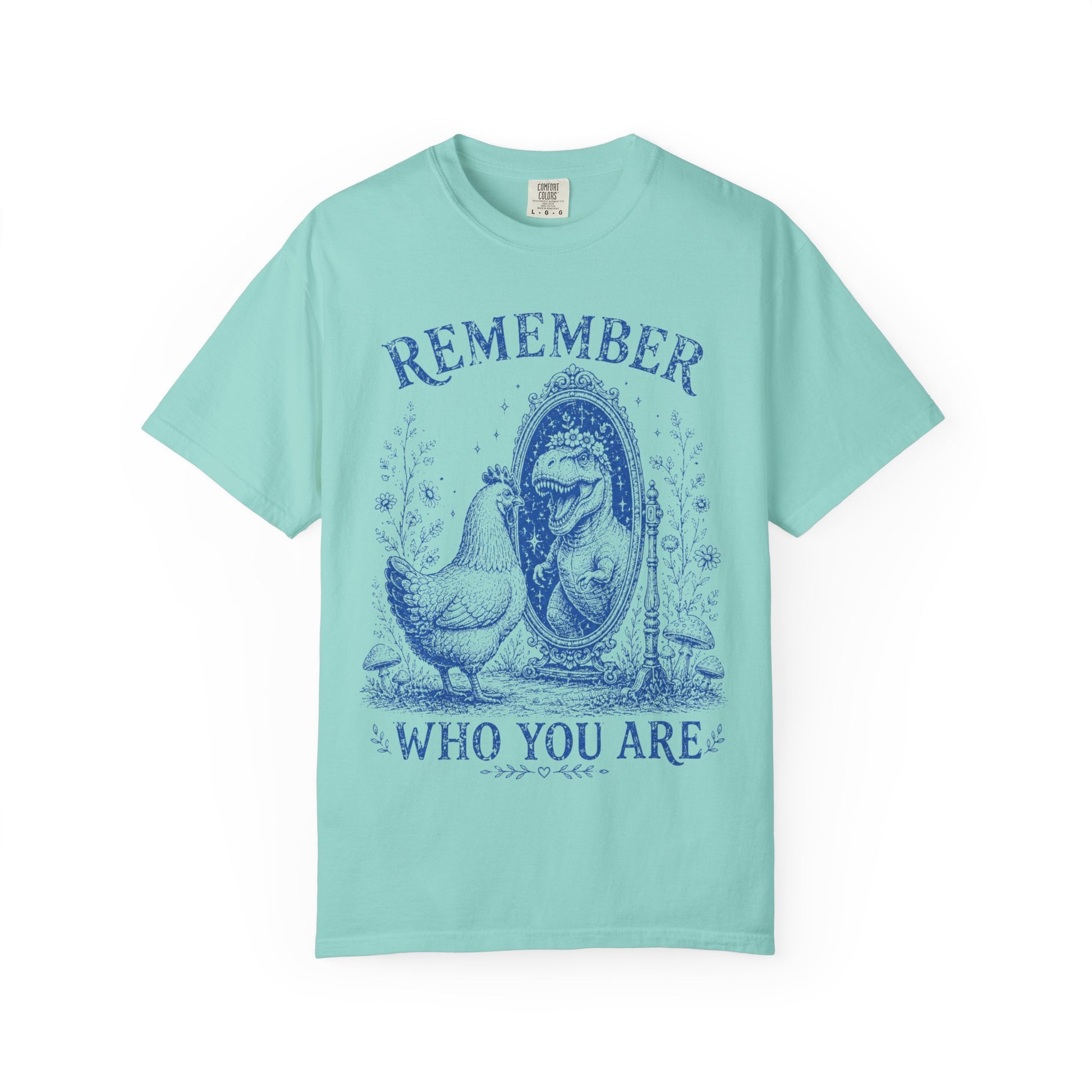 A light mint green short-sleeve t-shirt with a dark blue graphic print on the front. The graphic features a hen standing next to a mirror with a woman's face inside, with the words 'REMEMBER' above and 'WHO YOU ARE' below.