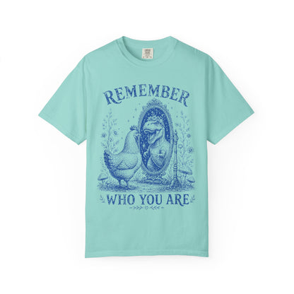 A light mint green short-sleeve t-shirt with a dark blue graphic print on the front. The graphic features a hen standing next to a mirror with a woman's face inside, with the words 'REMEMBER' above and 'WHO YOU ARE' below.