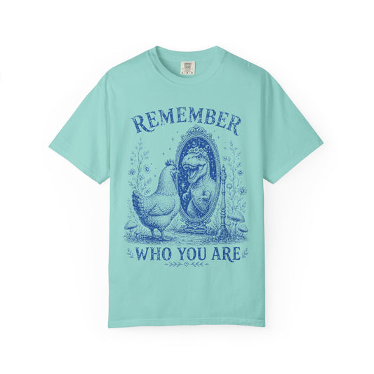 A light mint green short-sleeve t-shirt with a dark blue graphic print on the front. The graphic features a hen standing next to a mirror with a woman's face inside, with the words 'REMEMBER' above and 'WHO YOU ARE' below.